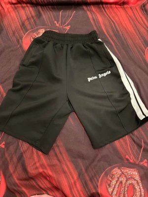 Palm Angels svarta shorts med vita ränder - Svarta shorts från Palm Angels med vita sidoremsor och logga tryckt på vänster ben. Modellen har elastisk midja med snörning för skön passform och är gjorda i ett mjukt syntetmaterial. Perfekta för en sportig och trendig look.