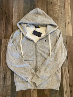 Grå hoodie från Polo Ralph Lauren - Snygg grå hoodie från Polo Ralph Lauren med dragkedja och vit snörning i huvan. Tröjan har det klassiska blå Polo-logot på bröstet och är fodrad med mjukt material på insidan. Perfekt för chill dagar och enkel att matcha med jeans eller joggers.