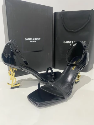 Svarta Saint Laurent sandaler med YSL-klack - Exklusiva svarta sandaler från Saint Laurent med glansig finish i skinn. Snygg fyrkantig tå och tunn rem över foten. Klacken är i guld och formad som YSL-loggan, vilket ger en lyxig vibe. Justerbart spänne vid ankeln för perfekt passform.