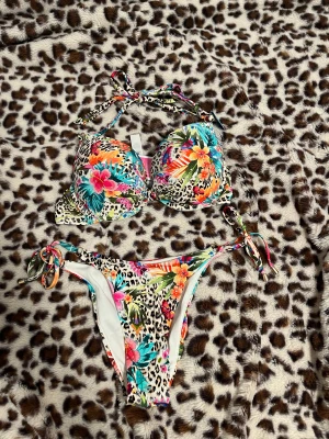 Leopardmönstrad blommig bikini XS/C - Färgstark bikini från New yorker med leopardmönster och blommor i rosa, orange, blå och grönt. Push-up överdel med knyt i nacken och dekorativa stenar. Underdel med snörning i sidorna. Perfekt för strandhäng och poolparty!