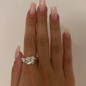 Silvrig ring med knutdesign - Säljer en snygg silvrig ring med en unik knutdesign framtill. Ringen har dubbla band som möts i en flätad, snurrad knut som ger en cool och modern vibe. Perfekt för dig som gillar statement-smycken och vill sticka ut lite extra.