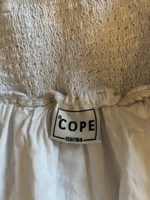 Vit kort topp från COPE - Supersöt vit kort topp från COPE med smockad överdel och justerbara smala axelband. Nedre delen av toppen är lätt rynkad och har ett diskret rutigt strukturmönster. Perfekt för dig som gillar en fräsch och enkel stil med lite volym upptill.