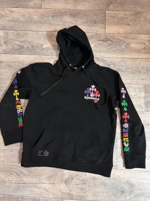 Svart Chrome Hearts hoodie med färgglada kors - Svart hoodie från Chrome Hearts med huva och magficka. Tröjan har färgglada kors i rött, grönt, blått, gult, rosa, vitt och lila på både rygg, bröst och ärmar.  Hoodie har jag fått som present och har ej något bevis på köp