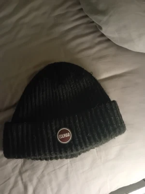 Svart ribbad mössa från Colmar - Snygg svart mössa från Colmar med ribbad stickning och uppvikt kant. Märkets logga sitter framtill i form av en rund patch. Perfekt för dig som vill ha en stilren och enkel look under kyliga dagar.                                                                               Ny pris 700 kr mitt pris 150 kr.                                        Skick 7/10