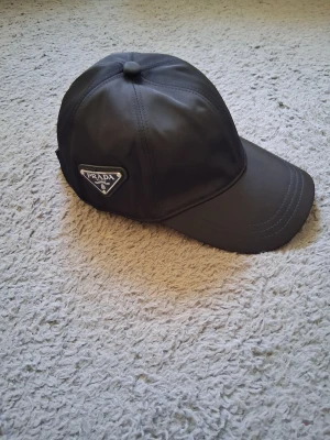 Svart keps från Prada i nylon - Snygg svart keps från Prada med klassisk böjd skärm och ikonisk metalltriangel-logga på sidan. Tillverkad i återvunnen nylon (Re-Nylon) med stilrena sömmar och justerbar passform. Perfekt för dig som gillar exklusiva accessoarer med street-vibe.