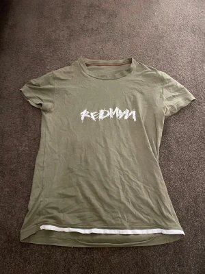 Olivgrön t-shirt från Hugo boss limiterad - Snygg olivgrön t-shirt från Hugo med coolt vitt tryck 'REDMYM' på bröstet och röd broderad Hugo-logga på ärmen. T-shirten har rund hals, korta ärmar och en vit detalj längst ner. Perfekt för dig som gillar streetwear och vill sticka ut.