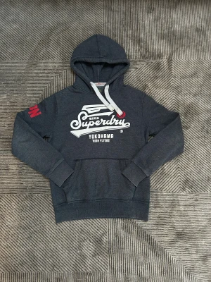Mörkgrå Superdry hoodie  - Snygg mörkgrå hoodie från Superdry med vit logga och text på bröstet samt röd detalj på ärmen. Tröjan har huva med vita snören och en stor magficka. Perfekt för chill dagar eller när du vill ha en sportig look.