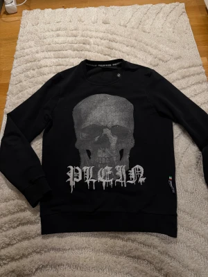 Philipp Plein tröja (Limited Edition) - Svart långärmad tröja från Philipp Plein med stor silverfärgad döskalle och logotyp på framsidan. Tröjan har ribbade muddar och en exklusiv PP-logga i metall på ryggen. Materialet är mjuk bomull och passformen är normal. Perfekt för dig som gillar edgy streetwear.