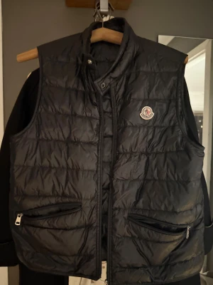Svart dunväst från Moncler - Snygg svart dunväst från Moncler med quiltad design och hög krage. Västen har två praktiska fickor med dragkedja framtill och Moncler-logga på bröstet. Perfekt att slänga över en hoodie för extra värme och stil.