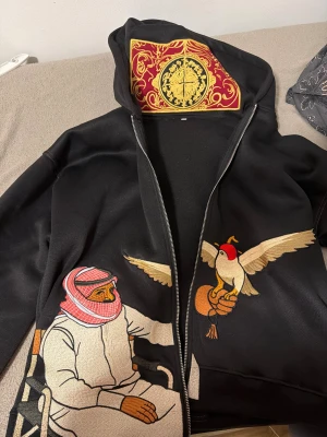 Svart hoodie med broderad konst - Unik svart hoodie med dragkedja och huva, dekorerad med broderade motiv av en man i traditionell klädsel och en fågel. Insidan av huvan har ett färgstarkt mönster i rött, gult och svart. Perfekt för dig som gillar statement-plagg och detaljerad design.