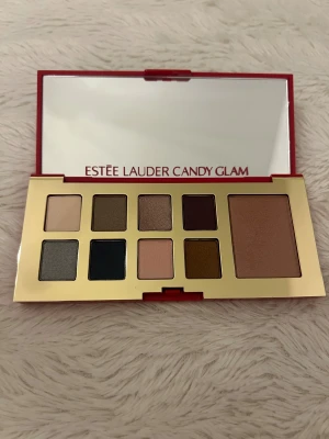 Estée Lauder Candy Glam palett - Snygg sminkpalett från Estée Lauder Candy Glam med 8 ögonskuggor i nyanser som beige, brun, guld, rosa, grå och mörkblå samt en rouge i varm rosabrun ton. Paletten har röd och guldig förpackning med stor spegel. Perfekt för dig som gillar att experimentera med färg och shimmer.