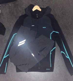 Svart hoodie från BOSS med blå detaljer - Svart hoodie från BOSS med snygga blå linjer längs ärmarna och BOSS-logga på bröstet. Tröjan har huva med snörning och dragkedja framtill. Perfekt för träning eller chill, med sportig vibe och modern design.