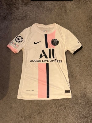 PSG Messi 2021/22 borta tröja player edition - Paris Saint-Germain fotbollströja med Messi 30-tryck, Champions League-märke på ärmen och klassisk PSG-logga. Tröjan är vit med en rosa och svart vertikal rand, kortärmad och tillverkad i lätt Dri-FIT-material. Perfekt för dig som älskar fotboll och Messi.