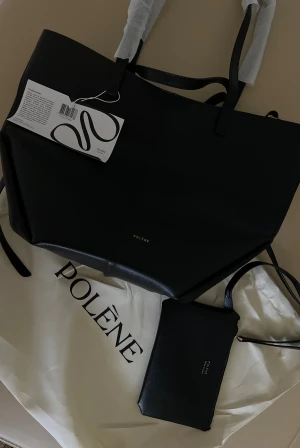 Svart väska från Polène - Stilren svart väska från Polène , minimalistisk design. Väskan har två långa handtag, magnetlås och medföljande svart clutch med dragkedja. Perfekt för dig som gillar klassiska och snygga accessoarer.