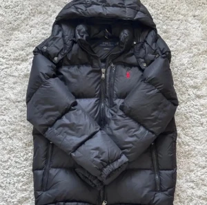 Svart dunjacka från Polo Ralph Lauren - Svart dunjacka från Polo Ralph Lauren med röd broderad logga på bröstet. Jackan har huva, flera dragkedjefickor och quiltad design. Materialet är syntet och dun, perfekt för kalla dagar. Stängs med dragkedja och har långärmade ärmar.