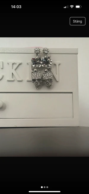 Glittriga döskalle-örhängen i silver - Unika örhängen med stora glittriga döskallar och stenar i silverfärg. Hängande design med flera detaljer som pärlor och facetterade kulor. Perfekt för dig som vill sticka ut med en edgy och cool stil. Säljs som ett par.