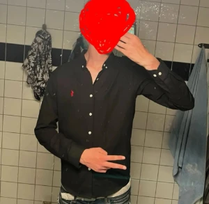 Svart skjorta från Ralph Lauren - Svart skjorta från Ralph Lauren med klassisk röd logga broderad på bröstet. Skjortan har vita knappar hela vägen och en snygg button-down krage. Långärmad och tillverkad i mjuk bomull, perfekt för en clean och stilren look.
