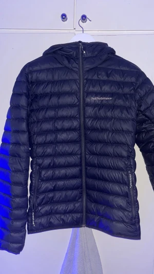 Svart dunjacka Peak Performance - Snygg svart dunjacka från Peak Performance med huva och dragkedja framtill. Jackan har quiltad design, två sidofickor med dragkedja och logga på bröstet. Perfekt för kyliga dagar och har en clean, sportig look.