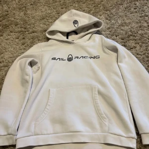 Vit hoodie från Sail Racing - Vit hoodie från Sail Racing med svart logotyptryck på bröstet och en mindre logga på huvan. Tröjan har en stor magficka och mjukt material som känns skönt mot huden. Klassisk passform med långa ärmar och ribbade muddar. Perfekt för chill dagar.
