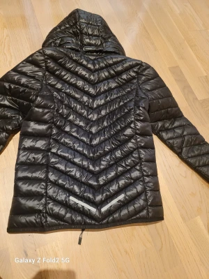 Svart dunjacka från McKinley - Säljer en svart dunjacka från McKinley med huva och snyggt quiltat mönster. Jackan har dragkedja framtill och reflekterande detaljer nedtill på ryggen. Perfekt för kalla dagar och riktigt skön att ha på sig.