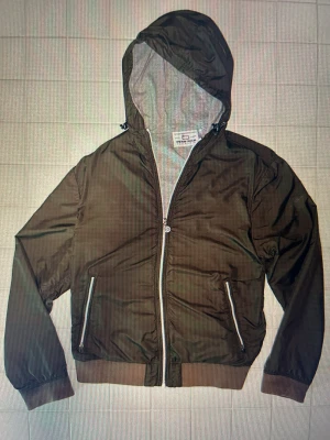 Woolrich Jacka - Snygg olivgrön vindjacka från Woolrich med grått foder i luvan. Jackan har dragkedja framtill och på fickorna, ribbade muddar och en loose passform. Perfekt med huva och stilrena detaljer i silver. Passar dig som gillar enkel och clean stil. Storlek XL i JR men sitter som en M i herrstorlek 