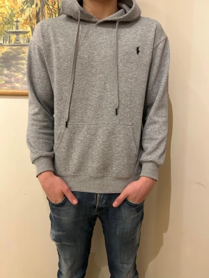 Grå hoodie från Polo Ralph Lauren - Snygg grå hoodie från Polo Ralph Lauren med klassisk logga broderad på bröstet. Tröjan har huva med snörning, känguruficka och ribbade muddar. Tillverkad i mjuk bomullsmix som är perfekt för chill dagar.