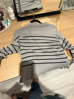 Grå randig stickad tröja - Säljer en grå stickad tröja med svarta horisontella ränder. Tröjan har ribbade muddar och nederkant samt lång ärm. Perfekt för dig som gillar en klassisk och chill stil.