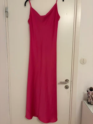 Rosa långklänning med tunna axelband - Säljer en snygg långklänning i stark rosa färg med tunna axelband och slinkig passform. Klänningen har en enkel, stilren design och faller mjukt längs kroppen. Perfekt för dig som gillar minimalistisk stil och vill sticka ut med färg.