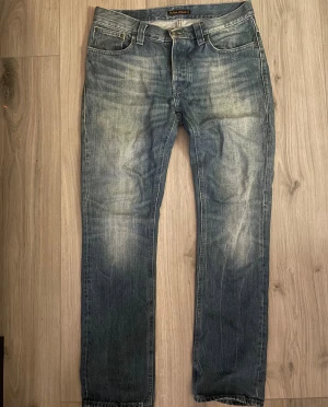 Nudie jeans grim tim  - Säljer ett par klassiska blå jeans från Nudie Jeans i storlek W32 L32. Jeansen har en straight fit, fem fickor och snygga slitningar framtill och baktill för en cool, avslappnad look. Materialet är robust denim och färgen är mellanblå med ljusare tvättade partier.