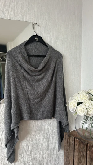 Grå stickad ponchotröja med draperad krage - Säljer en grå stickad ponchotröja med snyggt draperad krage och asymmetriska kanter. Perfekt för lager-på-lager och chill höststil. Mjuk och skön känsla, passar dig som gillar oversized och avslappnat.