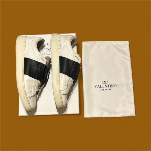 Valentino Garavani Open Sneakers 43 - Snygga Valentino Garavani Open sneakers i vitt med bred svart läderdetalj över sidan. Skorna har rund tå, platt sula och snörning. Tillverkade i mjukt kalvskinn för lyxig känsla. Kommer med original dustbag och låda. Perfekt för dig som gillar exklusiva sneakers.