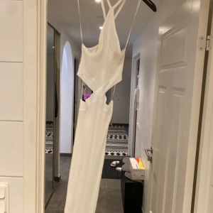 Vit ribbad långklänning med cut-outs - Säljer en vit ribbad långklänning med smala axelband och snygga cut-outs i midjan. Klänningen har omlott-detaljer och en figurnära passform som framhäver siluetten. Perfekt för dig som vill sticka ut med en trendig och stilren look.