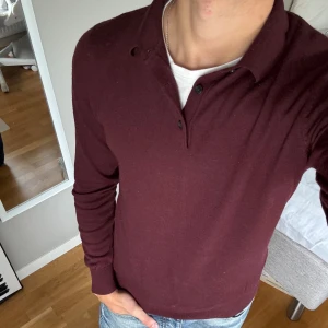 Vinröd pikétröja med lång ärm - Säljer en vinröd långärmad pikétröja med klassisk krage och knappar framtill. Tröjan har en slim passform och är tillverkad i ett mjukt material som känns skönt mot huden. Perfekt för dig som vill ha en stilren och enkel look.