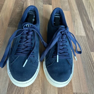 Mörkblå sneakers från Polo Ralph Lauren - Snygga mörkblå sneakers från Polo Ralph Lauren med klassisk logga på sidan och POLO-text bak. Skorna har rund tå, platt vit sula och snörning. Ovandelen är i syntetmaterial med textilfoder och gummisula. Perfekt för dig som gillar stilrena och sportiga sneakers.