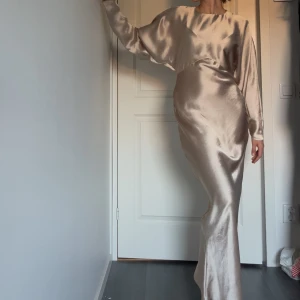 Champagnefärgad långklänning i satin - Elegant långklänning i skimrande champagnefärgad satin med lång ärm och rund halsringning. Perfekt för dig som vill sticka ut med en lyxig och modern look. Säljes pga att färgen inte passar mig.