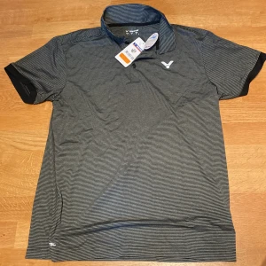 Victor - Randig grå pikétröja / badminton  - Snygg pikétröja från Victor i grått med tunna vita ränder. Tröjan har klassisk krage, korta ärmar och knäppning framtill. Materialet är mjukt och lätt polyester, perfekt för sport eller chill. Diskret logga på bröstet ger en clean look.