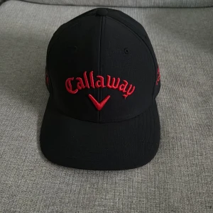 Svart Callaway keps med röd brodyr - Svart keps från Callaway med röd brodyr framtill och på sidorna, samt vit och röd Odyssey-logga bak. Klassisk form med böjd skärm och justerbar rem bak. Perfekt för golfbanan eller som sportig accessoar.