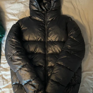 Svart pufferjacka från Grunt XXL - Säljer en svart pufferjacka från Grunt i storlek M. Jackan har en glansig finish, huva och dragkedja framtill. Perfekt för kalla dagar med sin rejäla vaddering och klassiska look. Enkel att matcha och riktigt skön att bära.