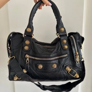 Svart Balenciaga City Bag med giant studs  - Balenciaga City i storleken ”Classic”! Svart mjukt läder med guldfärgade giant studs detaljer. En av Balenciagas mest ikoniska modeller med den perfekta kombinationen av vardagsväska och statement-piece. Lagom stor för att rymma det nödvändigaste men även en laptop.   Dubbla handtag och avtagbar axelrem gör att den kan bäras på flera sätt. Dragkedja upptill, frontficka med dragkedja och klassiska dekordetaljer. Spegel kommer med! Denna är köpt via vinted och därmed finns även en verifieringstag🤍