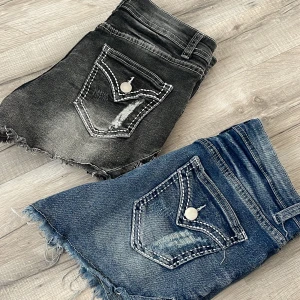 Två par jeansshorts med stjärndetaljer - Säljer två par snygga jeansshorts, ett par i klassisk blå denim och ett par i tvättad svart. Båda har råa kanter, slitningar och broderade vita stjärnor på sidorna. Bakfickorna har dekorativa sömmar och silverfärgade knappar. Perfekta för en cool och avslappnad stil.