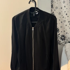 Svart bomberjacka från HM divided  - Svart bomberjacka från Hm Divided med klassisk ribbad krage och silverfärgad dragkedja framtill. Jackan har långärmade ärmar och två sidofickor. Materialet är lätt och har en diskret glansig finish som ger en cool vibe.