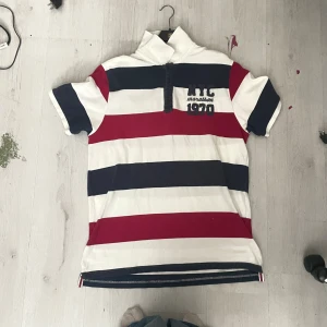 Randig pikétröja XXL från MyWear - Säljer en pikétröja från MyWear i storlek XXL med breda ränder i vitt, marinblått och rött. Tröjan har korta ärmar, krage och knappslå framtill. Broderad text 'NYC marathon 1970' på bröstet. Perfekt för dig som gillar sportig och avslappnad stil.