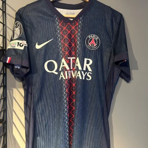 PSG matchtröja Nike herr S - Paris Saint-Germain fotbollströja i mörkblått med röda och vita detaljer, tillverkad i lätt och ventilerande material. Tröjan har korta ärmar, PSG-logga på bröstet, Qatar Airways-tryck och unikt mönster framtill. Nummer 14 och D.Doué på ryggen. Helt ny tröja med tagg en replika