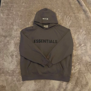 Grå Essentials Fear of God hoodie - Säljer en grön hoodie från Essentials Fear of God i storlek L. Tröjan har huva, stor svart logga på bröstet och diskret tryck på ärmen. Klassisk kanguruficka framtill och ribbade muddar. Perfekt för dig som gillar streetwear och enkel stil.