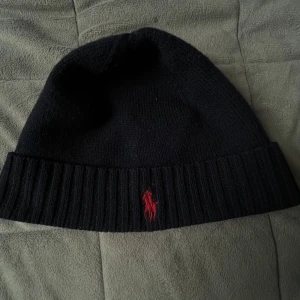 Svart merinoullsmössa från Polo Ralph Lauren - Svart mössa från Polo Ralph Lauren i mjuk merinoull. Klassisk ribbad kant och röd broderad Polo-logga framtill. Perfekt för kalla dagar och enkel att matcha med din stil. Snygg och tidlös design som funkar året runt.