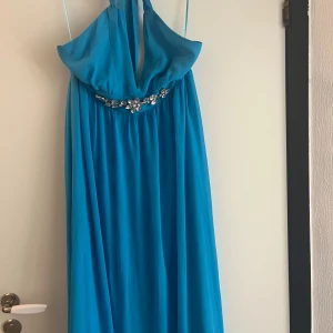 Blå långklänning med strassdetalj - Snygg blå långklänning med djup v-ringning och halterneck. Klänningen har en glittrig strassdetalj under bysten som ger extra bling. Tyget är lätt och faller fint, perfekt för speciella tillfällen. Stängs med dragkedja i ryggen.