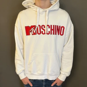 Vit hoodie Moschino x MTV - Vit hoodie från Moschino med stor röd MTV-logga och Moschino-tryck på bröstet. Tröjan har huva med snörning, magficka och ribbade muddar. Perfekt för dig som gillar streetwear och vill sticka ut med en fet logga.