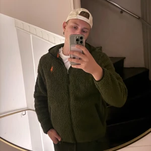 Grön teddyjacka från Polo Ralph Lauren - Mysig grön teddyjacka från Polo Ralph Lauren med dragkedja och orange logga på bröstet. Jackan har huva och är helfodrad med mjukt teddy-material, perfekt för kyliga dagar. Snygga svarta detaljer vid fickor och dragkedja. Passar dig som gillar streetwear och vill hålla värmen.
