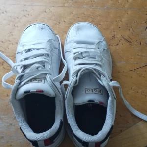 Vita sneakers Polo Ralph Lauren - Snygga vita sneakers från Polo Ralph Lauren med klassisk logga på sidan och färgdetaljer i rött, blått och vitt på hälen. Skorna har snörning, platt sula och är tillverkade i skinn med textildetaljer. Perfekta för en stilren och sportig look. Köpta för 1600. Priset kan diskuteras lägre. Skriv gärna innan ni köper!