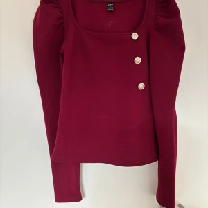 Vinröd topp med dekorativa knappar - Snygg vinröd topp från Shein med fyrkantig ringning och tre dekorativa vita knappar framtill. Långärmad modell med lätt puff vid axlarna. Perfekt för dig som vill ha en stilren men ändå unik look.